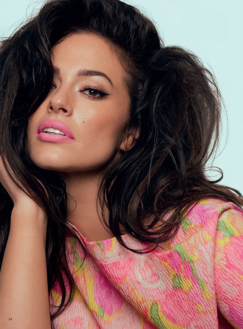 Close-up portrait of an adult female model with dark wavy hair and pink lipstick wearing a floral patterned top in a photo studio.