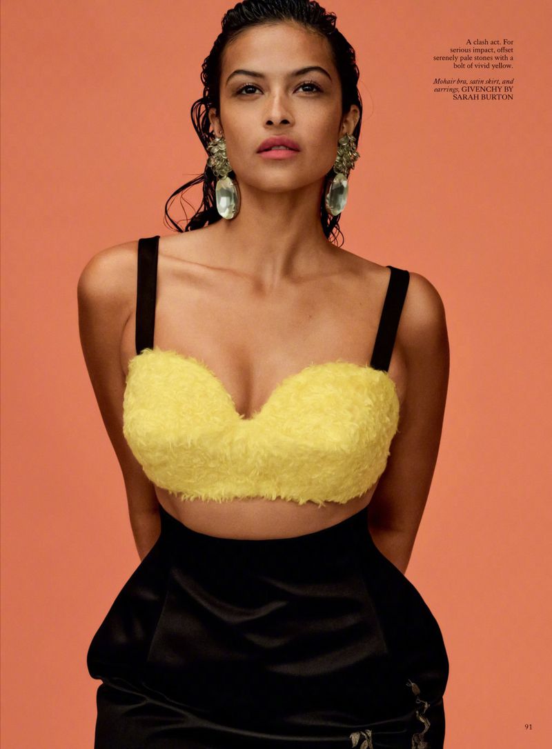 Portrait of a woman with wet hair wearing a yellow top and black skirt, posing against a peach-colored background.