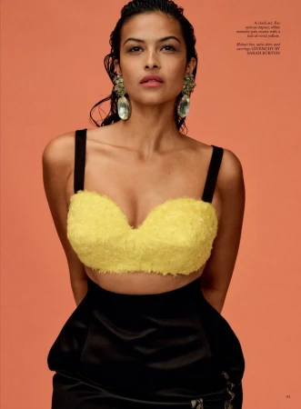 Portrait of a woman with wet hair wearing a yellow top and black skirt, posing against a peach-colored background.
