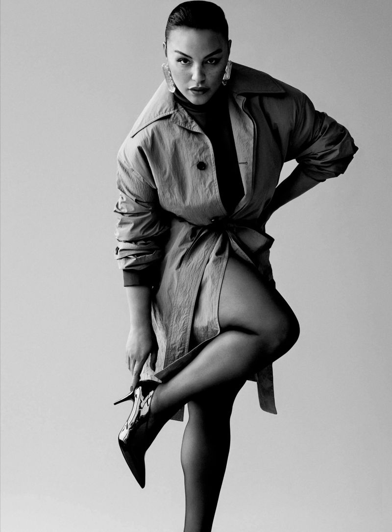 Black-and-white fashion portrait of a woman in a trench coat and high heels, posing against a neutral background in a photo studio.