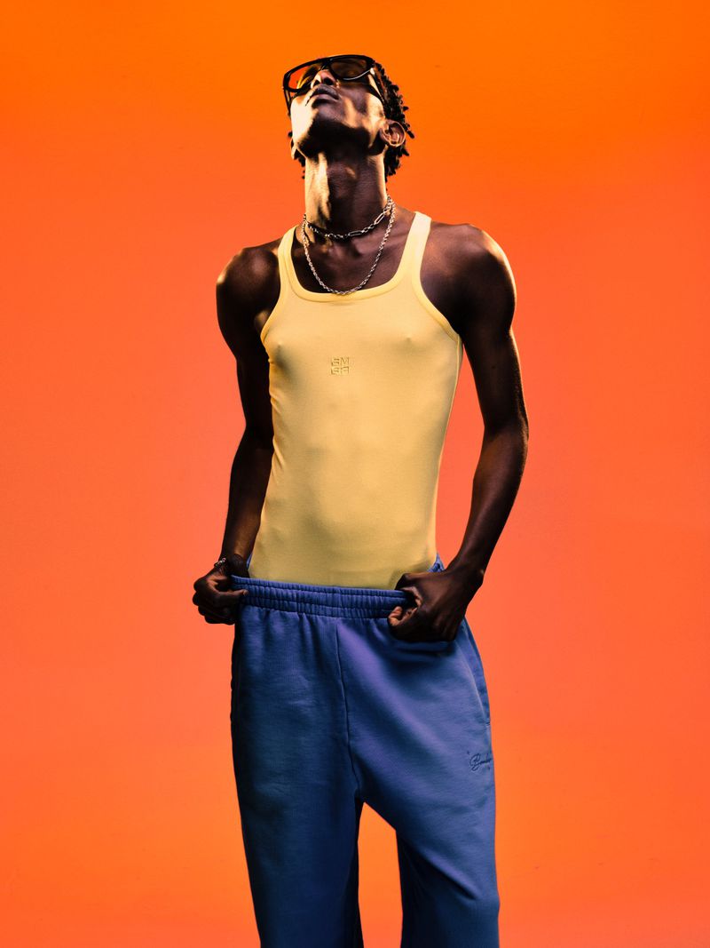 Model wearing a yellow tank top and blue sweatpants against a vibrant orange backdrop