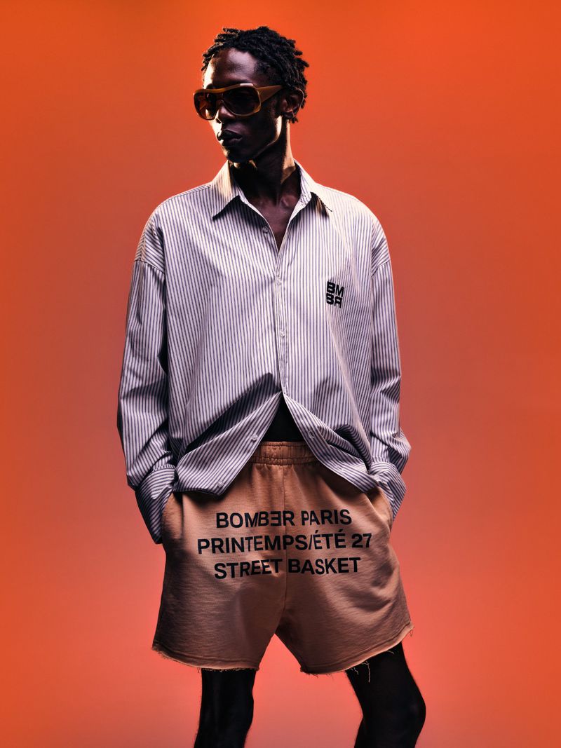 Black model wearing striped shirt, tan shorts and sunglasses against an orange backdrop for the Bomber spring summer 27 collection