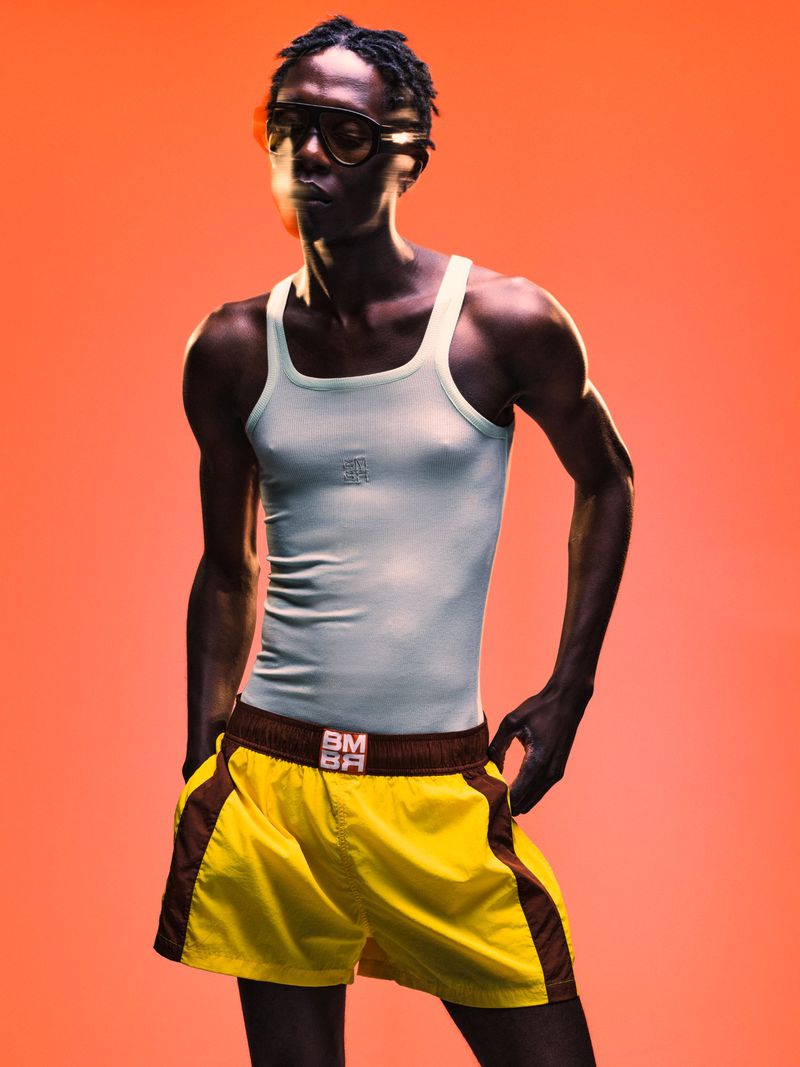 Black man wearing dark sunglasses, white tank top, and yellow shorts against an orange studio background