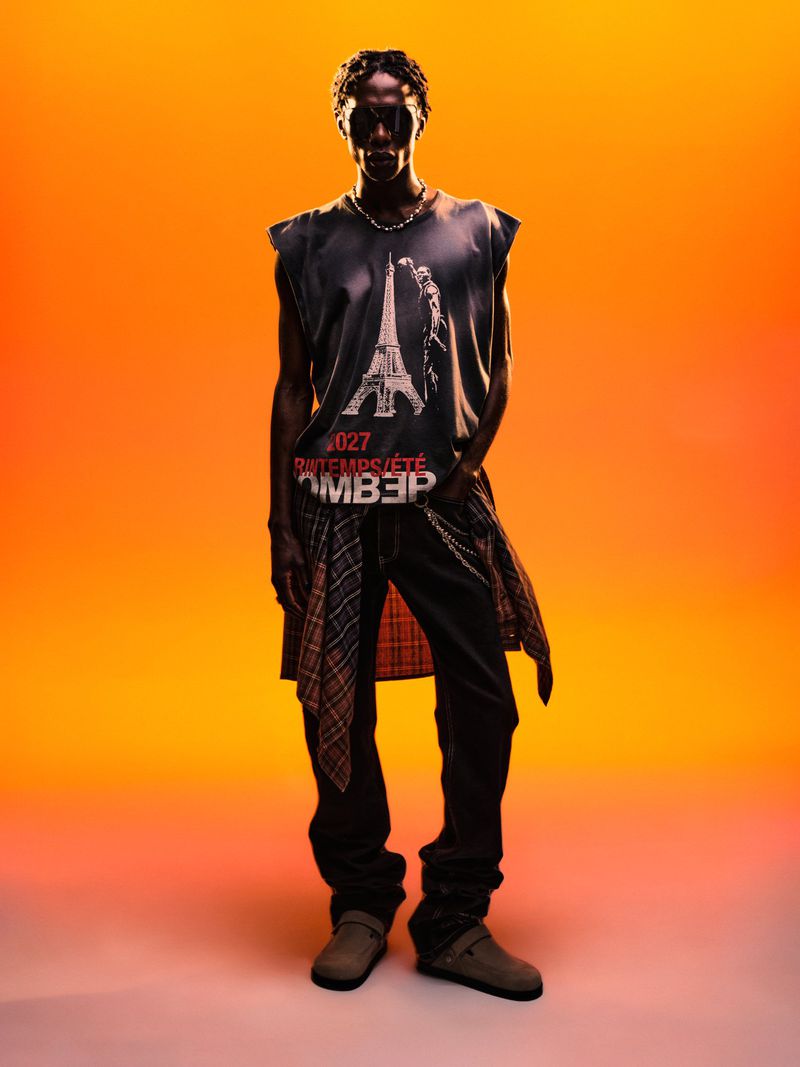 Black man wearing graphic print sleeveless top, dark trousers, and flannel shirt tied around waist against orange gradient backdrop