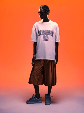 Model wearing a graphic t-shirt and wide shorts posing against a warm orange gradient background for Bomber spring summer 27.