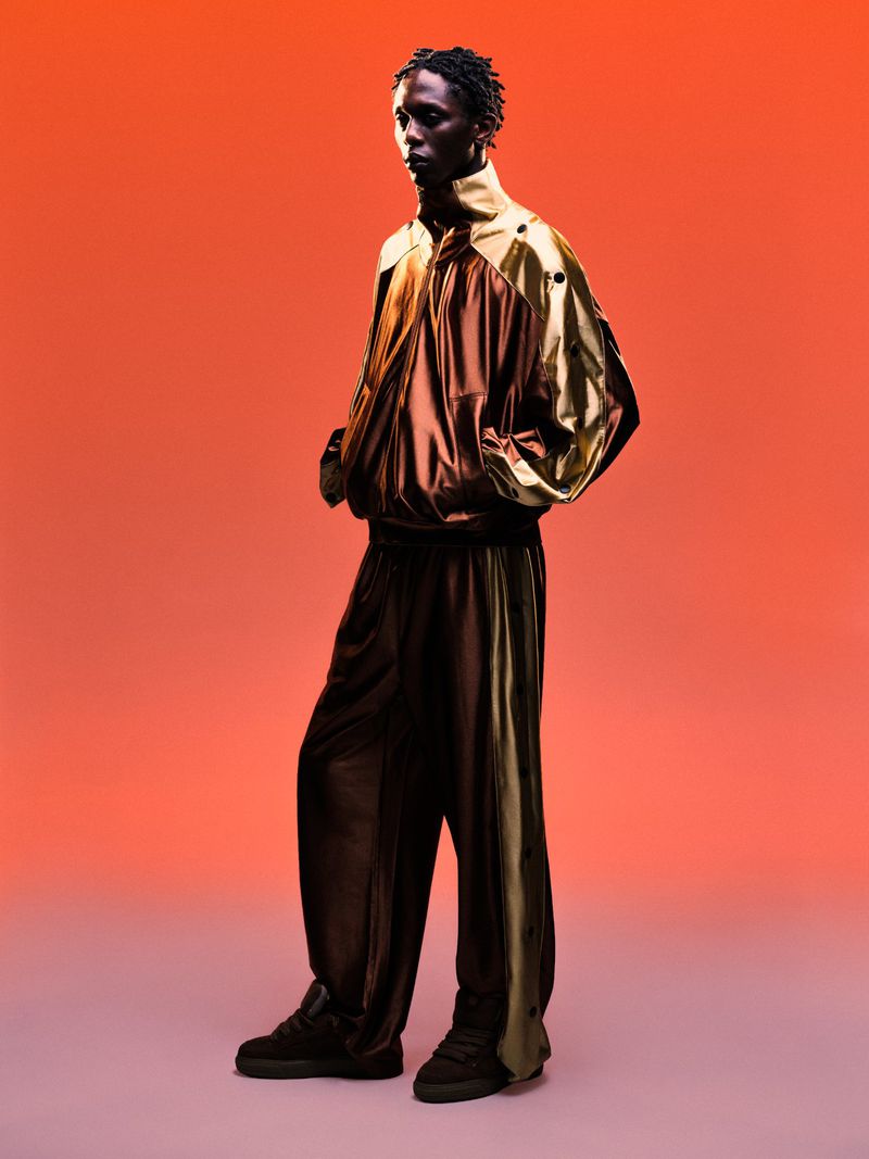Model Marcel Mbowole wearing a metallic gold and brown track jacket and matching trousers against an orange gradient background.