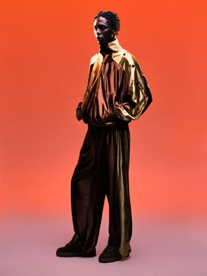 Model Marcel Mbowole wearing a metallic gold and brown track jacket and matching trousers against an orange gradient background.
