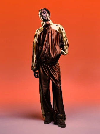 Model Marcel Mbowole wearing a glossy brown and gold track suit against a warm orange gradient background