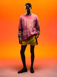 Model Marcel Mbowole wearing a pink graphic t-shirt and yellow shorts against a warm orange gradient background.