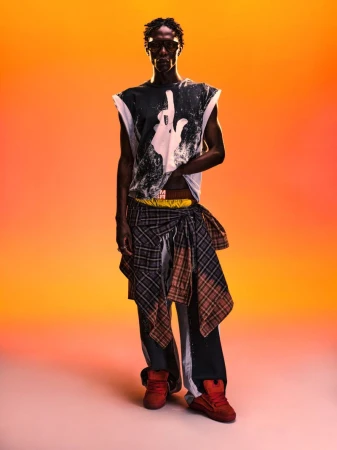 Black man wearing sunglasses, graphic sleeveless top, layered plaid shirt tied at waist, and red sneakers against an orange gradient background
