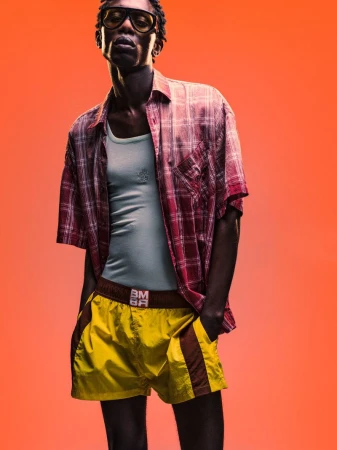 Model Marcel Mbowole wearing a plaid shirt and yellow shorts against an orange studio background