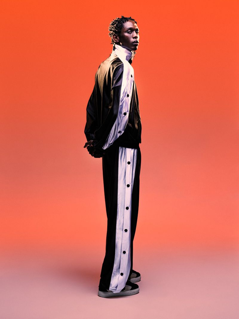 Black male model wearing a black jacket and matching trousers with white side panels against a vibrant red gradient backdrop.