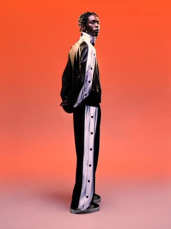 Black male model wearing a black jacket and matching trousers with white side panels against a vibrant red gradient backdrop.