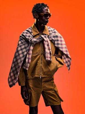 Model in brown zip jacket, matching shorts, plaid shirt draped over shoulders, and sunglasses against an orange background.