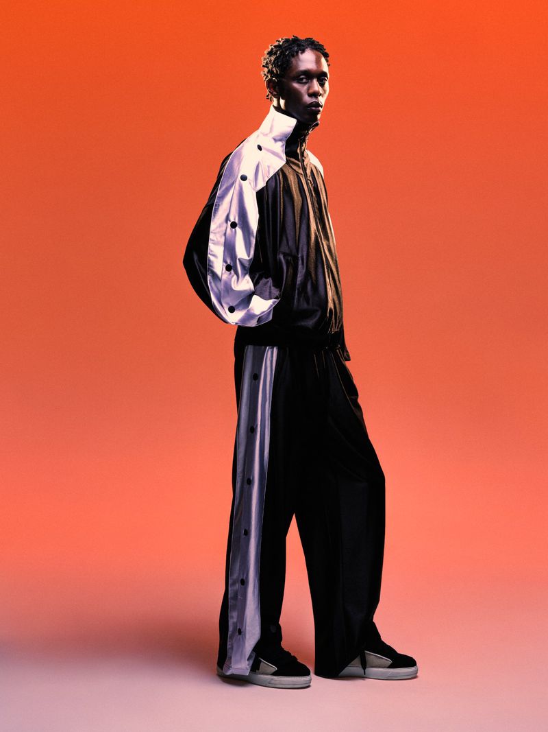 Model Marcel Mbowole wearing a black and white tracksuit against a warm gradient studio backdrop for a spring summer lookbook