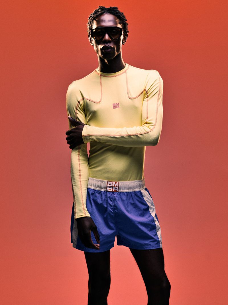 Model wearing yellow long-sleeve top and blue athletic shorts against a warm red-toned background