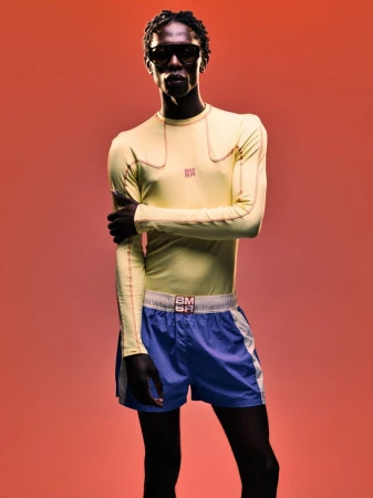 Model wearing yellow long-sleeve top and blue athletic shorts against a warm red-toned background