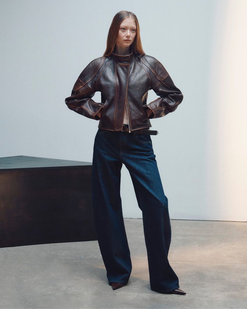 Model standing against a plain background wearing a dark brown leather jacket and wide blue jeans.