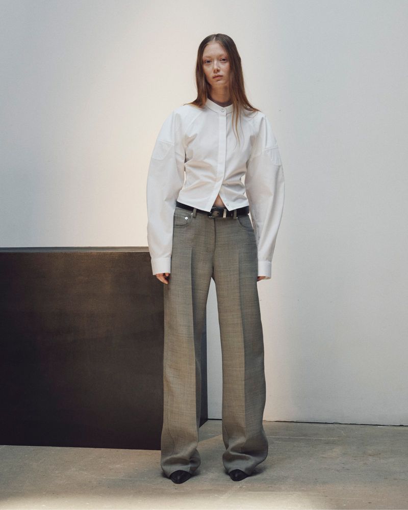 Model standing in a white button-down shirt and grey trousers against a minimalist studio wall
