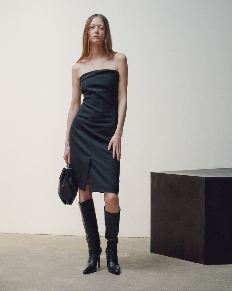 Model wearing a strapless black dress and knee-high boots holding a handbag against a plain background