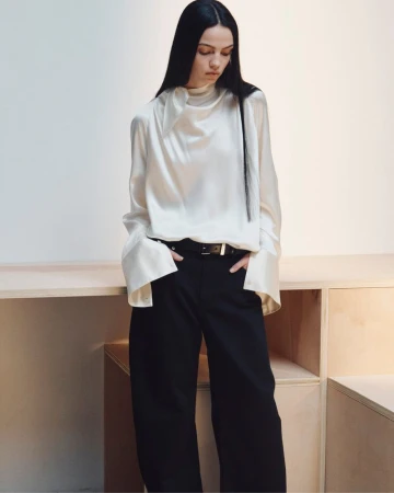 Model with long dark hair wearing a white silk blouse and black trousers posing indoors near wooden shelving