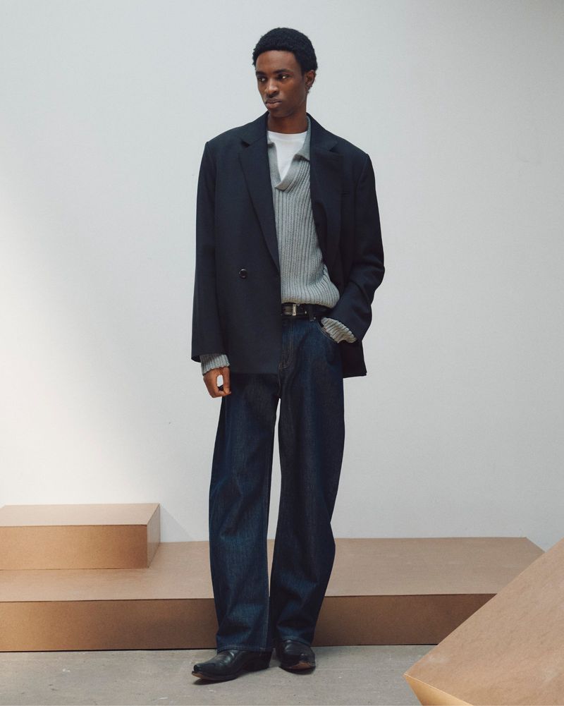 A man wearing an oversized black blazer, ribbed grey sweater, and dark denim trousers standing against a pale studio wall.