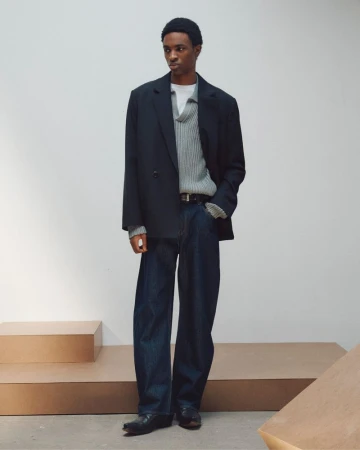 A man wearing an oversized black blazer, ribbed grey sweater, and dark denim trousers standing against a pale studio wall.