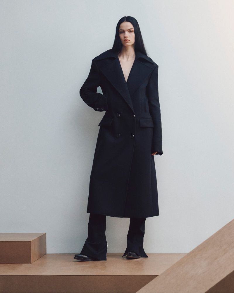 Model standing against a white seamless backdrop wearing a long black coat over trousers