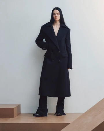 Model standing against a white seamless backdrop wearing a long black coat over trousers