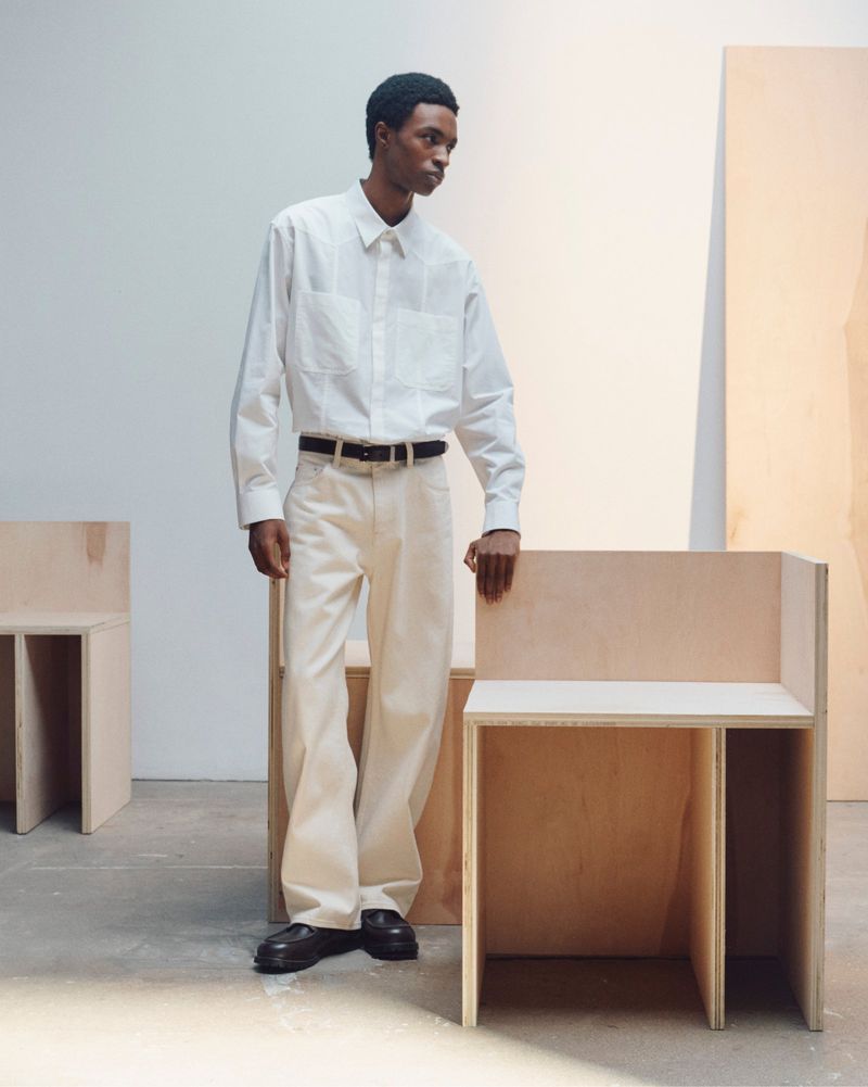Man wearing a white shirt and cream trousers standing in a studio with wooden furniture elements.