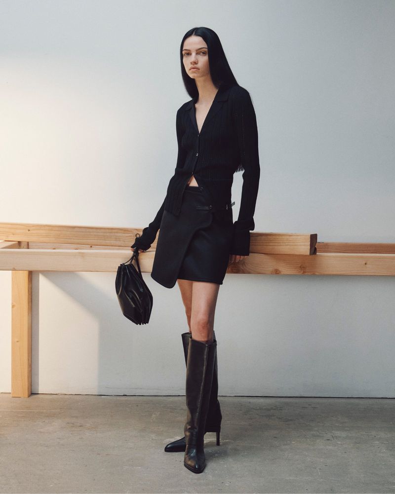 Model standing in black clothing with tall boots and a handbag against a plain wall with a wooden beam