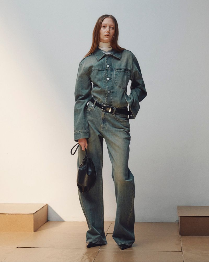 Model in full denim outfit wearing a matching jacket and wide trousers holding a black handbag against a light wall