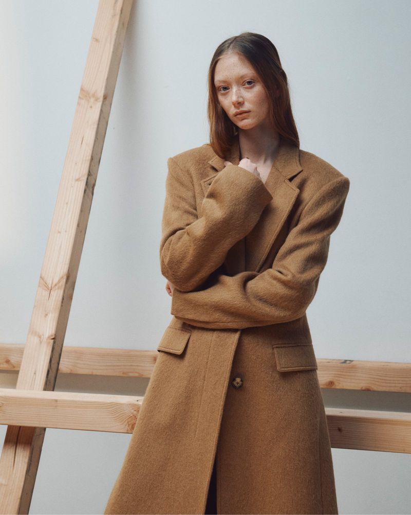Model standing against a plain white wall wearing a long camel wool coat near wooden beams