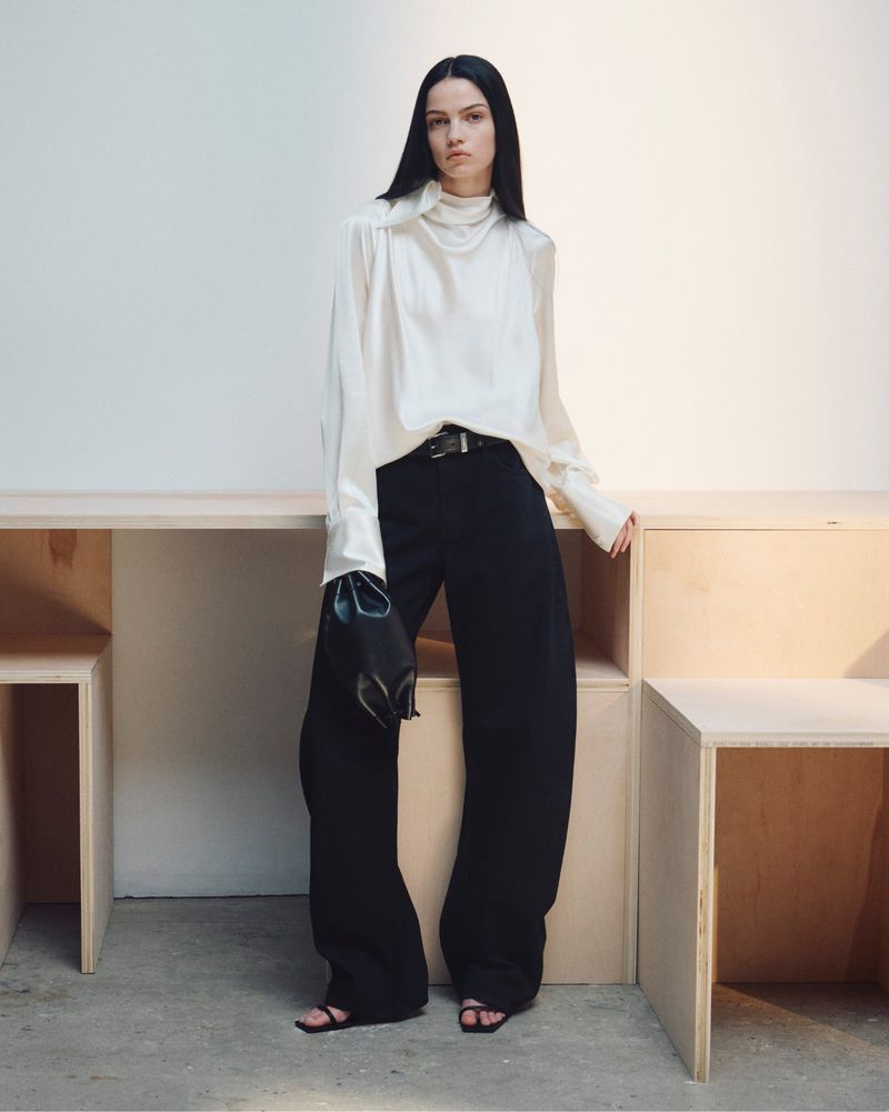 Model standing against modular wooden furniture wearing an ivory silk blouse and black trousers for Helmut Lang