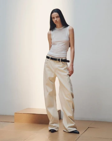 Model standing against a plain white wall wearing a white tank top and light trousers with a black belt