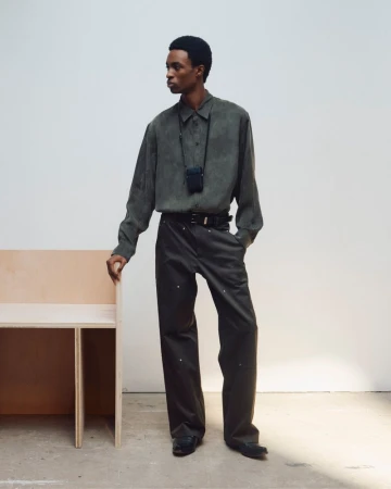 A male model standing in a muted green button-down shirt and dark trousers against a white backdrop with a wooden bench.