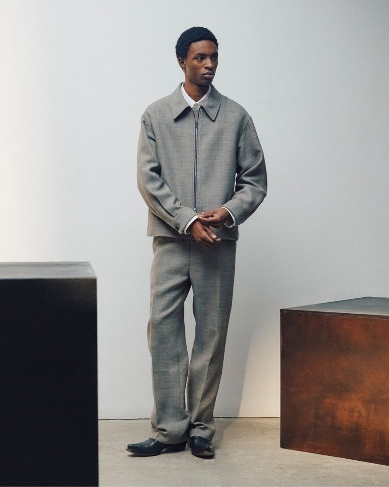 Full-length portrait of a male model wearing a grey wool zip-up jacket and matching trousers against a pale studio backdrop.