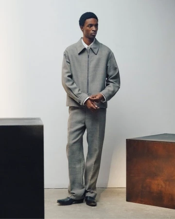 Full-length portrait of a male model wearing a grey wool zip-up jacket and matching trousers against a pale studio backdrop.