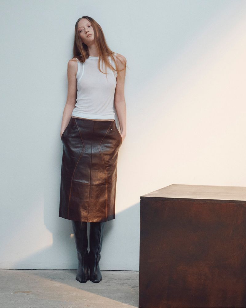 Model in white tank top and dark leather skirt leaning against white wall beside dark block