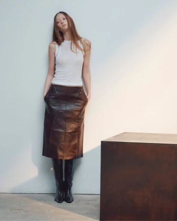 Model in white tank top and dark leather skirt leaning against white wall beside dark block
