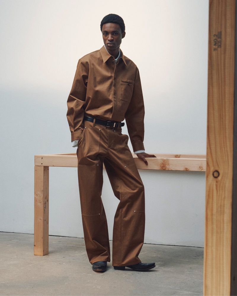 Black man wearing a brown shirt and trousers standing beside a wooden frame in a studio