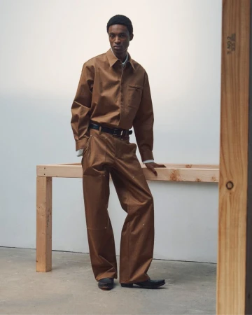 Black man wearing a brown shirt and trousers standing beside a wooden frame in a studio