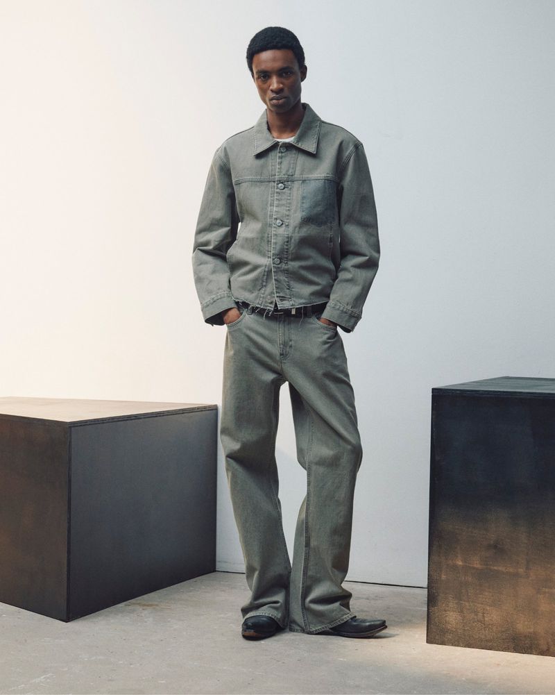Model wearing a grey denim jacket and matching jeans standing in a studio with dark geometric blocks.