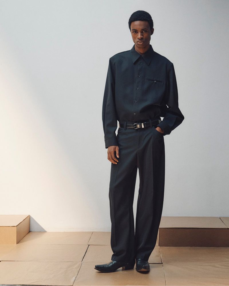 Man wearing dark button-down shirt, trousers, and beanie standing on wooden platform against a plain grey studio backdrop