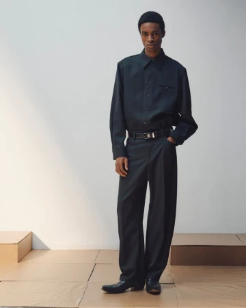 Man wearing dark button-down shirt, trousers, and beanie standing on wooden platform against a plain grey studio backdrop