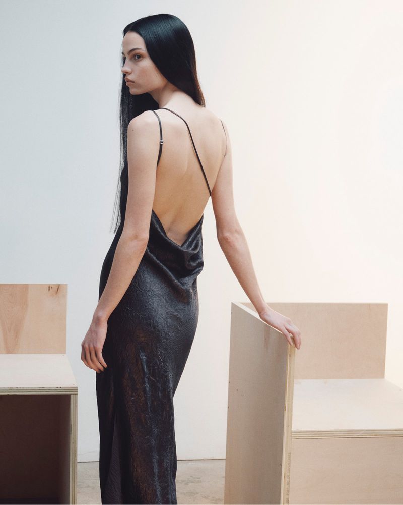 Model with long dark hair wearing a backless dark slip dress posing in a minimalist studio setting.