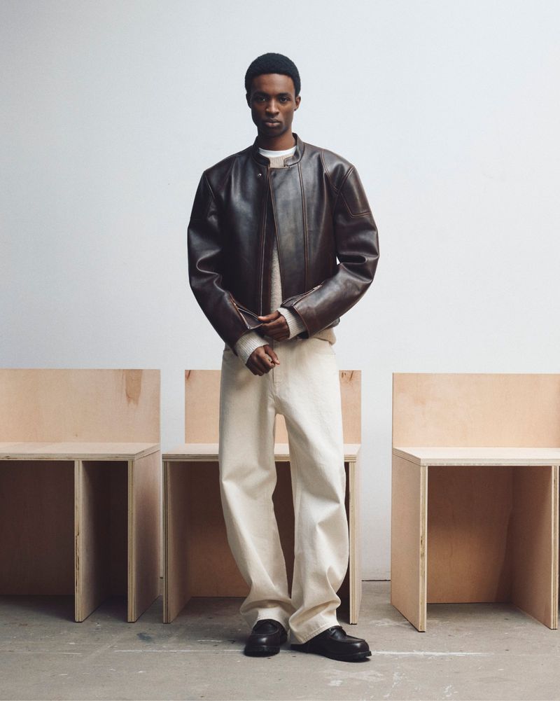 Model wearing a dark brown leather jacket and cream trousers standing before minimalist wooden block chairs against a plain white wall