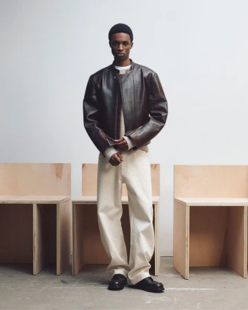 Model wearing a dark brown leather jacket and cream trousers standing before minimalist wooden block chairs against a plain white wall