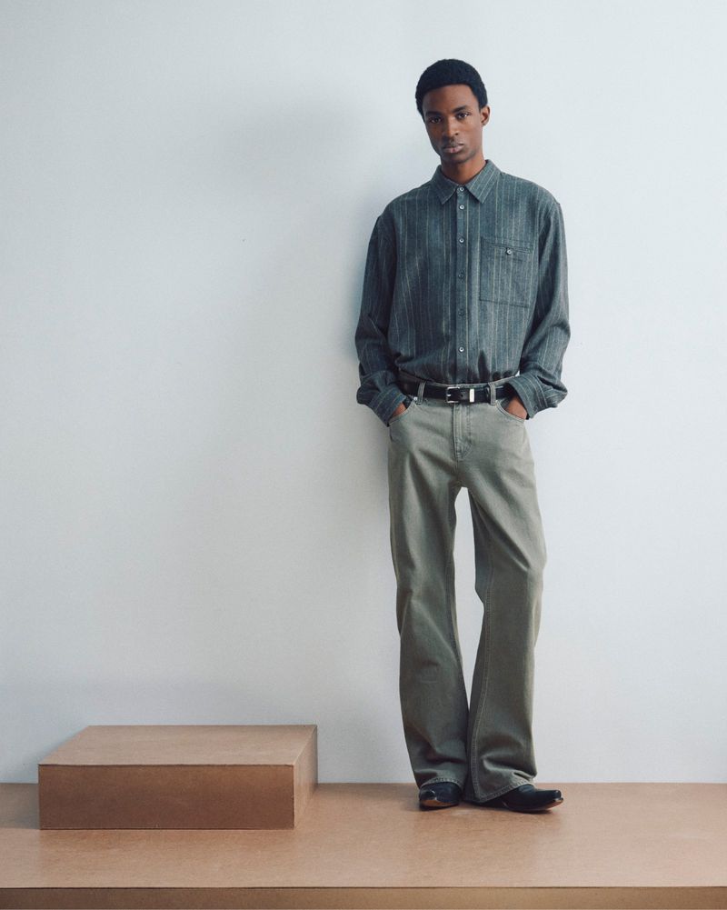 Model wearing a textured green shirt and grey trousers standing against a plain white studio background.