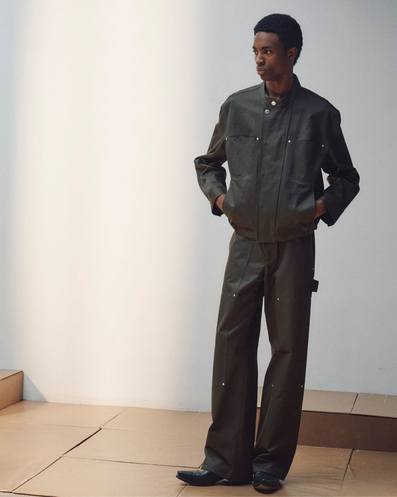 A man wearing a dark olive jacket and matching trousers standing against a light background.